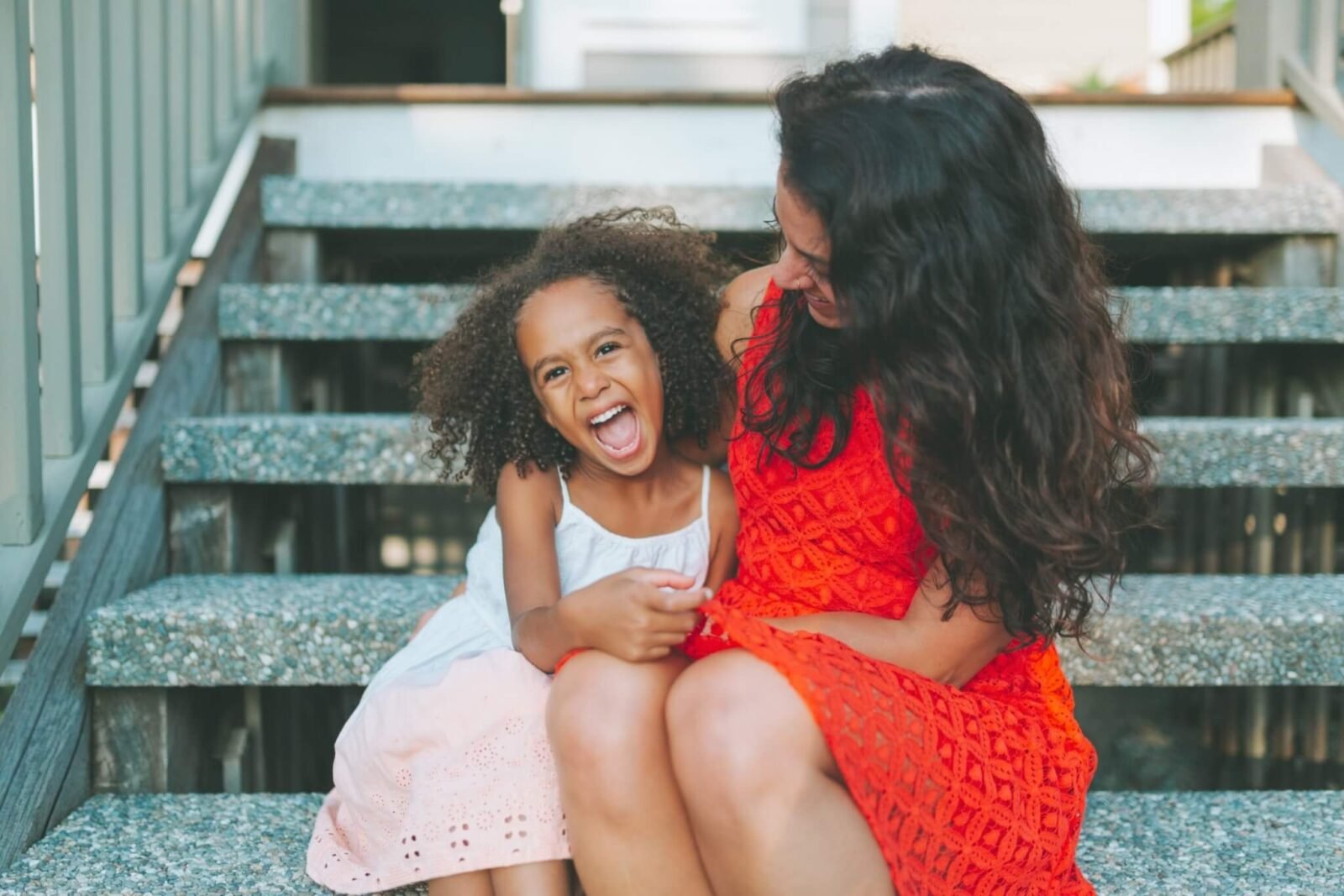 A Black woman is sitting on steps with a Black child. The woman is wearing a red dress. The child is wearing a white and pink dress. They are embracing and laughing.