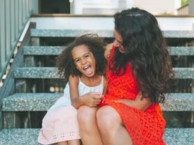 A Black woman is sitting on steps with a Black child. The woman is wearing a red dress. The child is wearing a white and pink dress. They are embracing and laughing.