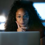This is an image of a Black woman looking at a laptop computer. She has glasses on and appears to be thinking.