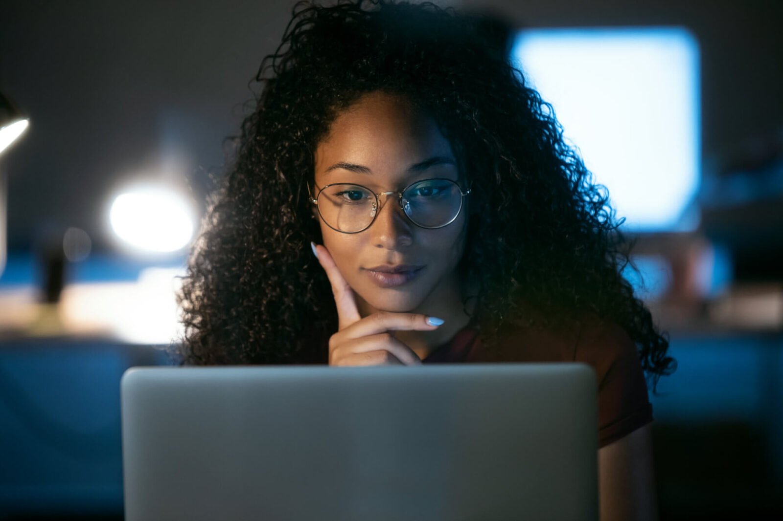 This is an image of a Black woman looking at a laptop computer. She has glasses on and appears to be thinking.