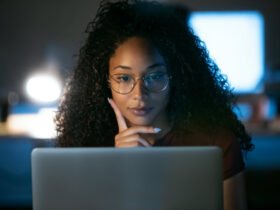 This is an image of a Black woman looking at a laptop computer. She has glasses on and appears to be thinking.