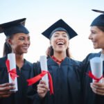 Three friends are seen post-college graduation talking and laughing with one another. They are wearing their caps and gowns and holding diplomas.