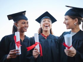Three friends are seen post-college graduation talking and laughing with one another. They are wearing their caps and gowns and holding diplomas.