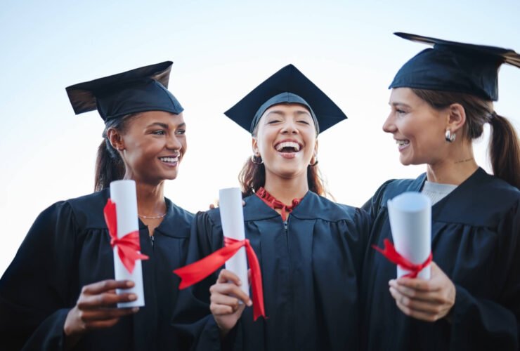 Three friends are seen post-college graduation talking and laughing with one another. They are wearing their caps and gowns and holding diplomas.