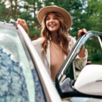 A woman is seen getting into the driver side of a white car. She has long auburn hair, is smiling, wearing a cream colored sweater, as well as a wide brimmed hat.