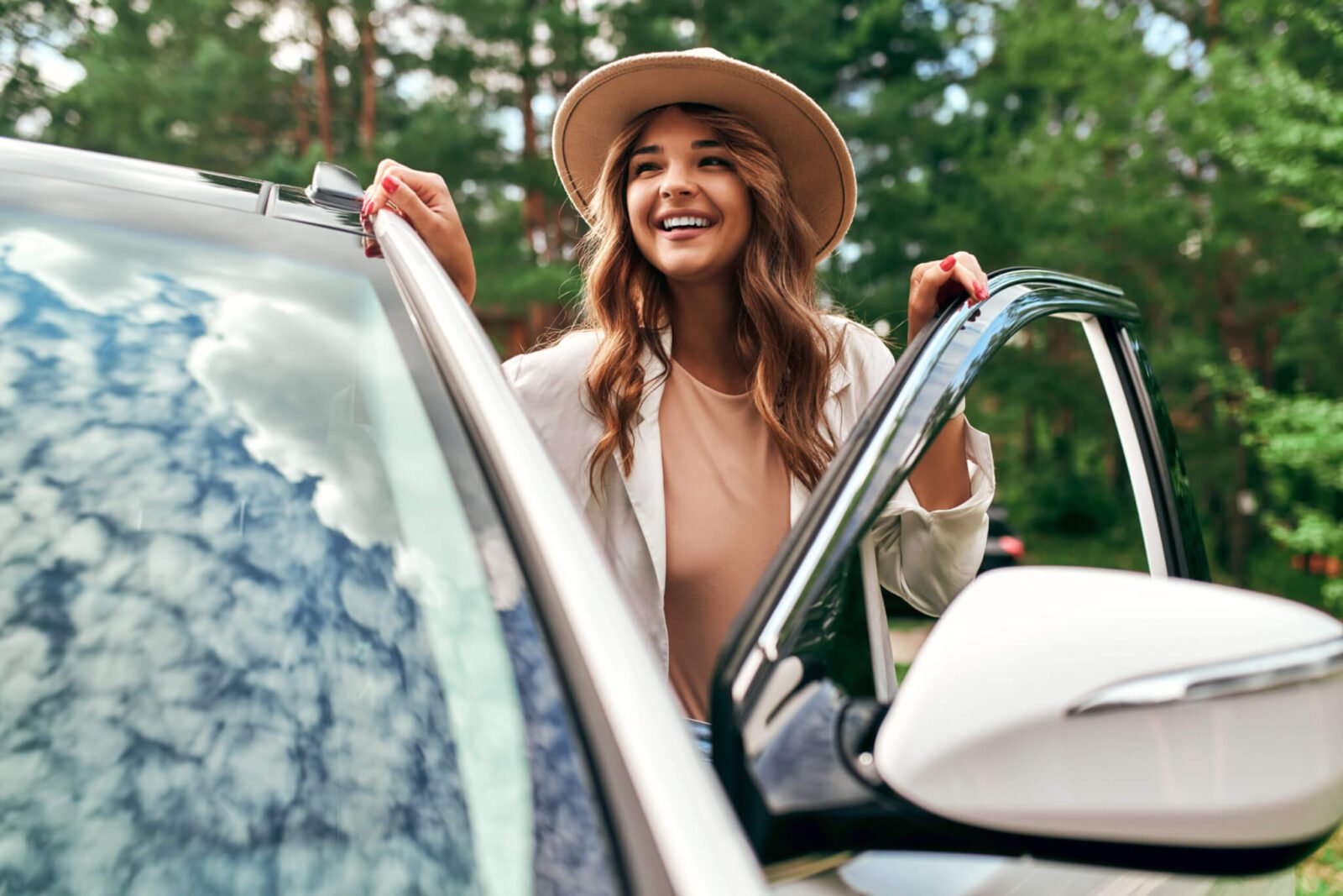A woman is seen getting into the driver side of a white car. She has long auburn hair, is smiling, wearing a cream colored sweater, as well as a wide brimmed hat.