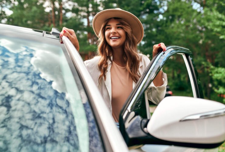 A woman is seen getting into the driver side of a white car. She has long auburn hair, is smiling, wearing a cream colored sweater, as well as a wide brimmed hat.
