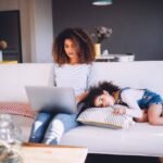 A mom sits on a couch typing on her laptop. Her daughter is laying beside her.