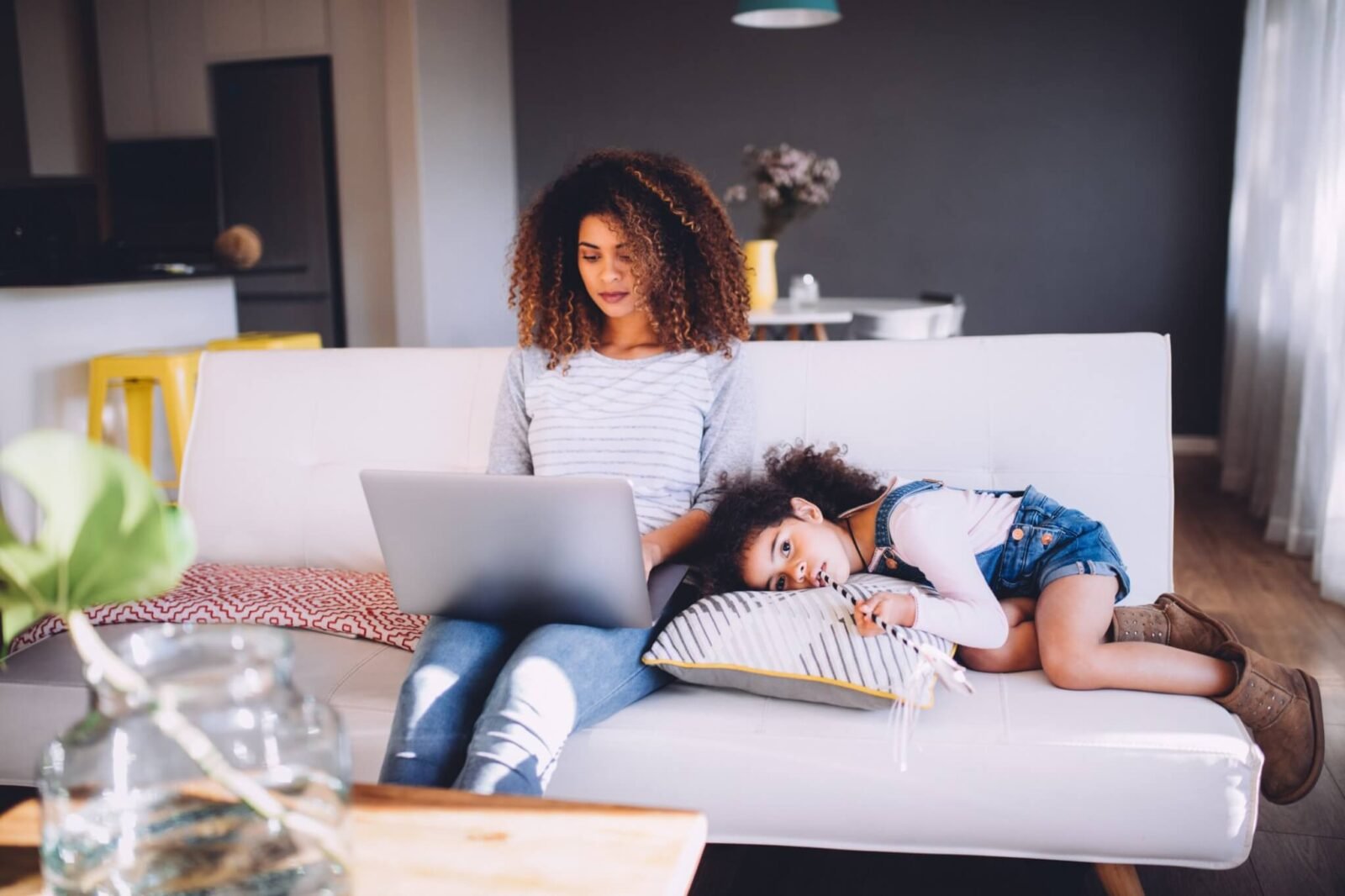A mom sits on a couch typing on her laptop. Her daughter is laying beside her.