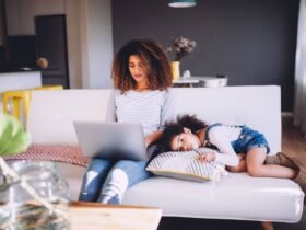 A mom sits on a couch typing on her laptop. Her daughter is laying beside her.