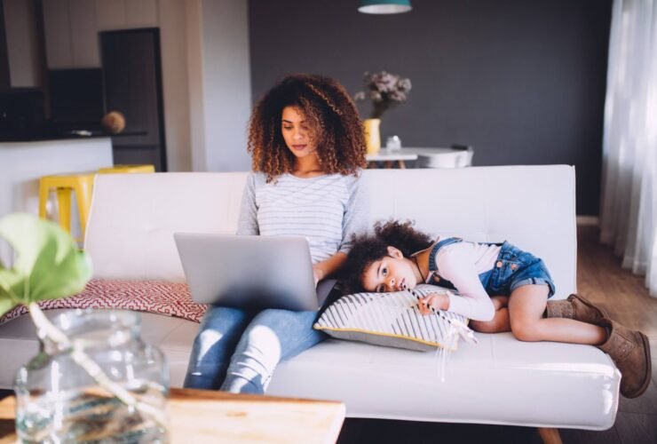A mom sits on a couch typing on her laptop. Her daughter is laying beside her.