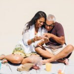 couple sitting on a beach with snacks smiling during their mini retirement