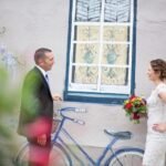 A couple looks at each other on their wedding day. They are standing outdoors in front of a window and bike.